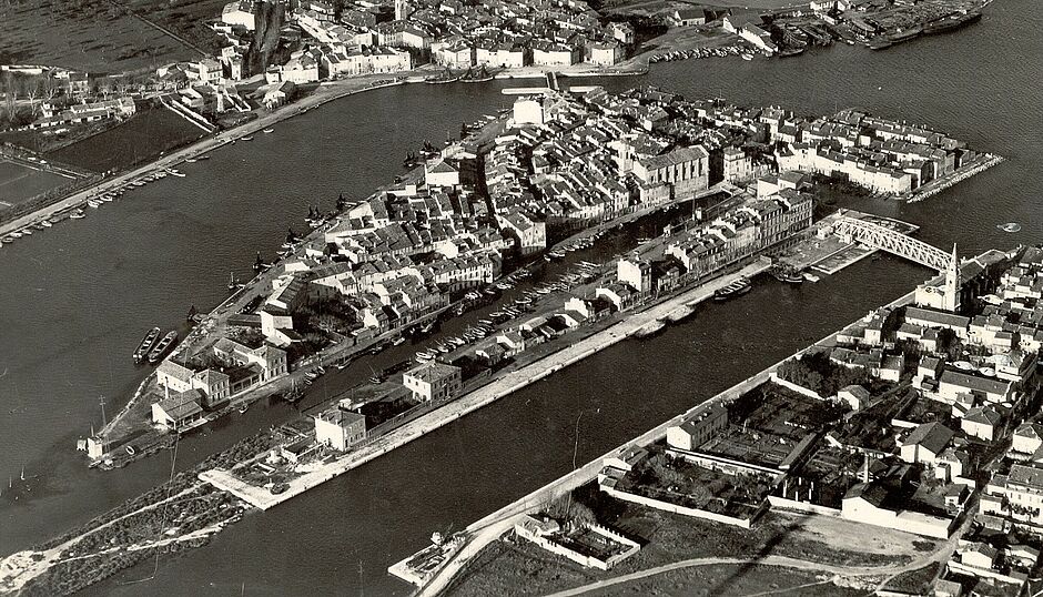 Vue aérienne du quartier de L’Île, entre 1930 et 1949 (détail). L’école de filles est à gauche de la pointe de L’Île, entre le canal Baussenque et le canal Saint Sébastien. À la gauche du bâtiment se trouve le tignadou, où étaient teints et pendus les filets de pêche. Sur le quai en face se situait encore l’école de garçons (à l’emplacement actuel de la médiathèque Louis Aragon) (crédits Archives Communales de Martigues) - Agrandir l'image (fenêtre modale)